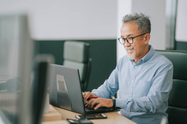 Asian chinese senior man with facial hair using laptop typing working in office open plan